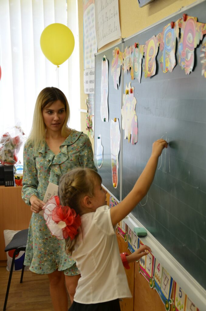 A teacher watching a young girl write on the blackboard